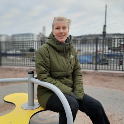 Kristina Salo, en kvinna med ljust bakåtkammat hår och olivgrön vinterjacka, sitter på en liten karusell för barn.