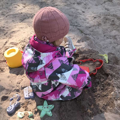 Ett litet barn i halare och mössa leker i en sandlåda.