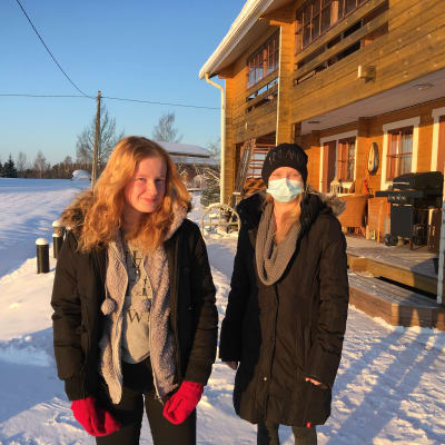 Tytär ja äiti seisovat talonsa edessä auringonpaisteessa talvella