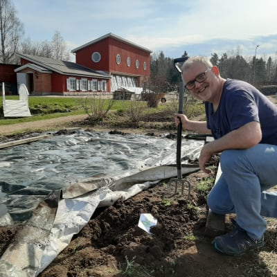 Man med grep sitter på knä vid ett trädgårdsland och ler in i kameran.