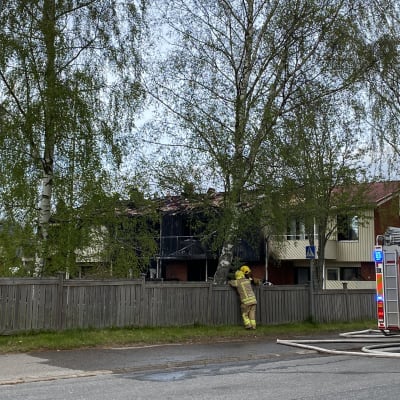 Ett tvåvåningsradhus som brunnit, av fyra lägenheter är de två mittersta förkolnade. På gatan står två brandmän och diskuterar. Bakändan av en brandbil syns.
