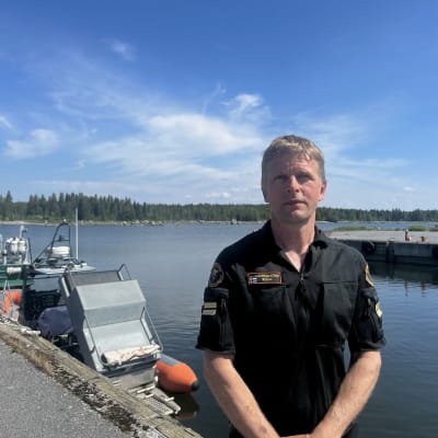Man i svart klädsel står på brygga vid havet. En båt syns i bakgrunden. 