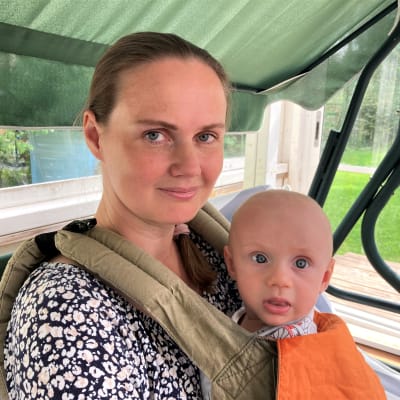 Nainen istuu puutarhakiikussa vauva kantorepussa.