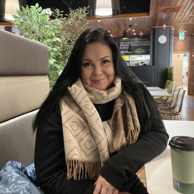 Laura Rasi istuu ravintolassa, katsoo kameraan ja hymyilee. Tummat hiukset,  vaalea huivi.