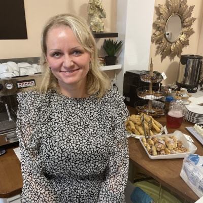 Vaaleahiuksinen nainen, yrittäjä Anastasia Lahti musta-valkoisessa mekossa kahvilan myyntitiskin edessä. Hymyilee kuvaajalle. Myyntitiskillä erilaisia leivonnaisia. 