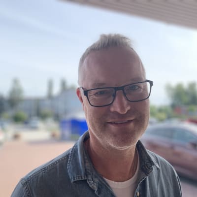 Mansperson med glasögon tittar in i kameran och ler. I bakgrunden bilar på parkeringsplats.