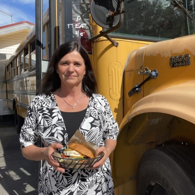 Bus Burger -ravintoloiden yrittäjä Eeva Laajoki seisoo keltaisen amerikkalaistyylisen koulubussin edessä ja pitelee käsissään metallisessa tarjoiluastiassa olevaa vuohenjuustohampurilaista.