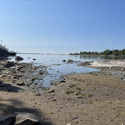 Kuvassa alhainen merenpinta Helsingin Ullanlinnassa Cafe Ursulan laiturin vieressä. Kuvassa hiekkaa, merta ja taustalla saari. Vasemmalla osa laiturista.