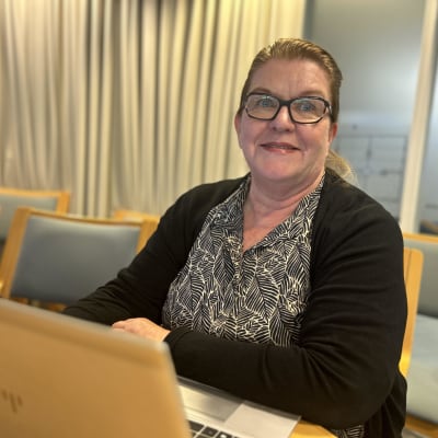 Silmälasipäinen nainen istuu neuvotteluhuoneessa läppärin ääressä ja katsoo kameraan.
