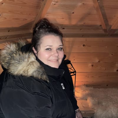 Hymyilevä nainen mustassa toppatakissa katsoo kohti kameraa. Tila on tunnelmallinen kota. 
