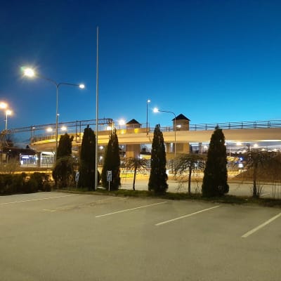 Asfalterade parkeringsplatser och gröna buskar framför en vit bro nattetid.