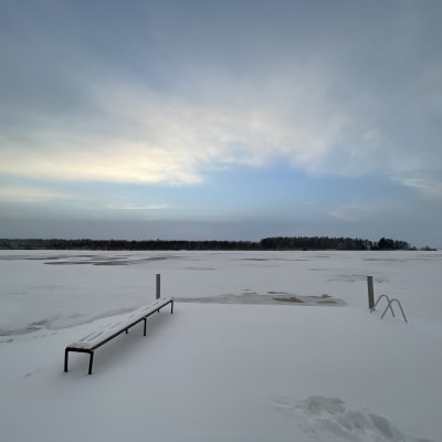 Järven rannalla oleva laituri peittyy lumeen. Taustalla näkyy tummia kohtia jäästä.