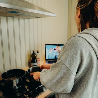 En kvinna står vid en kokspis med blicken vänd mot en dator som står bredvid. På skärmen syns en kvinna som skålar med ett glas vin.