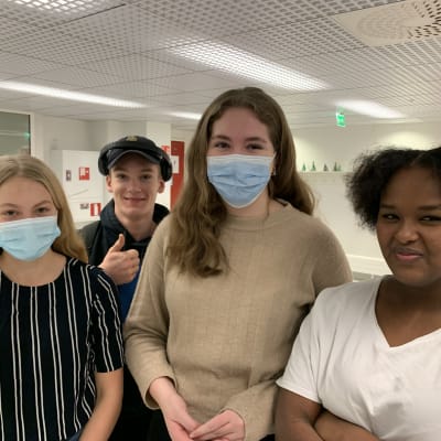 Fyra unga personer står intill varandra i skolmiljö, tittar in i kameran och ler. Två av dem bär munskydd.