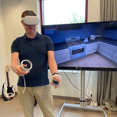 Kajaanin ammattikorkeakoulun pelituottaja Kyösti Koskela pelaa pakopeli Mysteeriä VR-lasit päässään. Kädessään hänellä on ohjaimet, joilla voi esimerkiksi noukkia lattialta roskia.