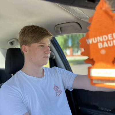Heikki Toivonen istuu autossaan.  Oranssi wunderbaum roikkuu kuvan etualalla. 