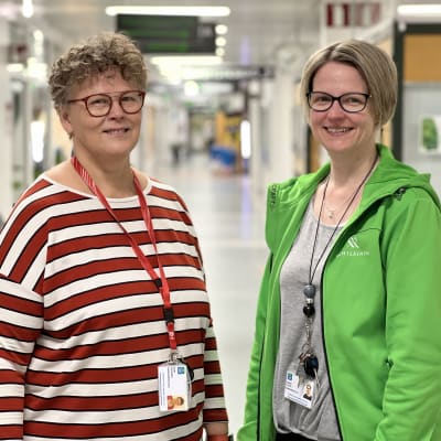 Kaksi naista seisoo sairaalan käytävällä ja katsoo kameraan. Oikeanpuoleisella naisella on yllään vihreä takki, jossa on teksti Mehiläinen.