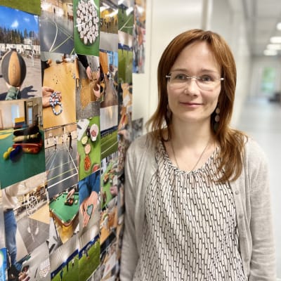 Nainen, jolla on silmälasit, vaaleanruskeat pitkät hiukset ja korvakorut nojaa seinään ja katsoo kameraa kohti. Hänen vieressään seinällä on valokuvia.