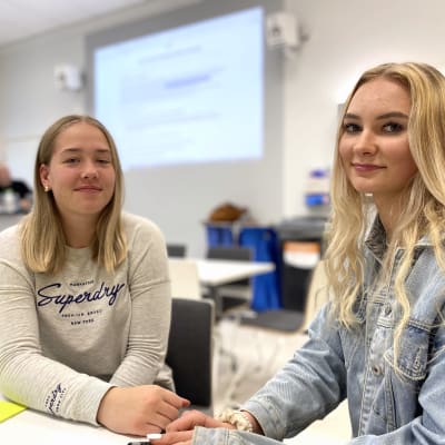 Kaksi vaaleatukkaista lukiolaistyttöä istuu pöydän ääressä ja katsoo kameraa kohti vienosti hymyillen.