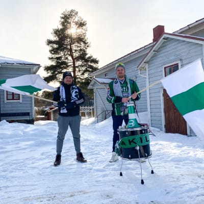Två personer står ute på en snöig gårdsplan och håller i grönvita flaggor. De har också en trumma och en megafon framför sig, och Akilles-tröjor och halsdukar.