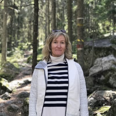 Blond kvinna ler framför kameran i skogsmiljö.