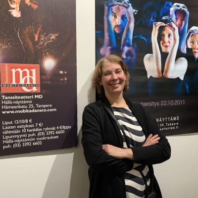 Nainen raidallisessa puserossa ja mustassa villatakissa (Tanssiteatteri MD:n toiminnanjohtaja Anniina Kumpuniemi) nojaa hymyillen seinään, jolla on tanssiteosten mainosjulisteita.