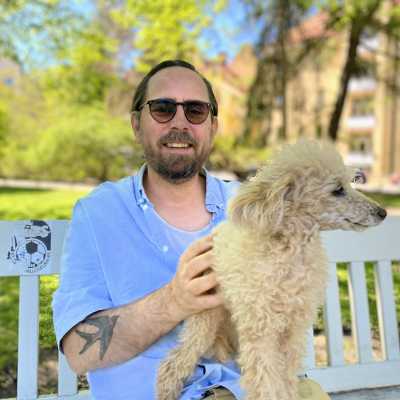 Philip Teir sitter i en blå skjorta med sin fluffiga hund i famnen i en park.