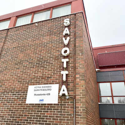 Bild på tegelhusfasad. På en skylt står det Kotka Svenska Barnträdgård. Bokstäver bildar ordet Savotta.