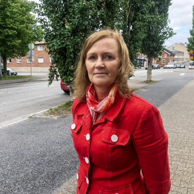 Kvinna i röd jacka står på trottoar. Bilar och träd i bakgrunden.
