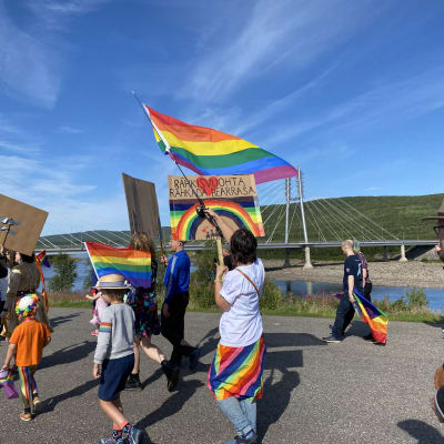 Sápmi Pride -kulkueessa Utsjoella heiluu sateenkaarilippuja ja kyltti, johon on kirjoitettu saamenkielinen teksti, joka tarkoittaa "rakkaus rakentaa perhettä".
