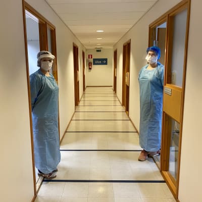 Två laboratorieskötare i skyddsdräkter står i var sin dörröppning i en korridor. 