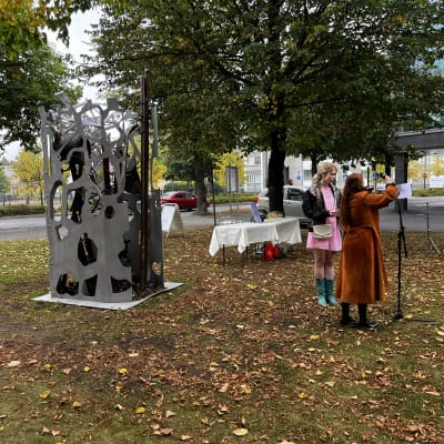 Metallinen patsas. Edessä kaksi naista valmisatutumassa puheeseen.
