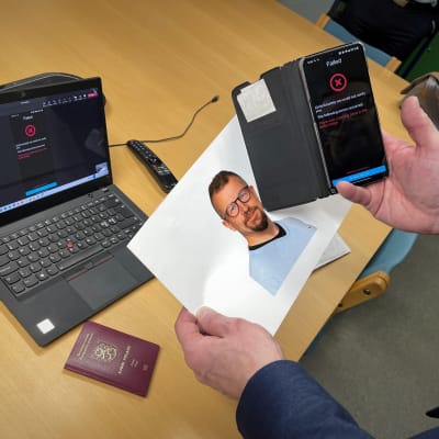 Kannettava tietokone pöydällä ja sen vieressä Suomen passi. Etualalla valokuva miehestä ja kännykästä kuvan päällä.