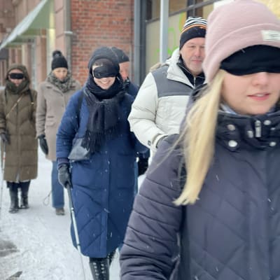 Ihmisiä kävelyllä lumisella kadulla laput silmillä, valkoisten keppien kanssa.