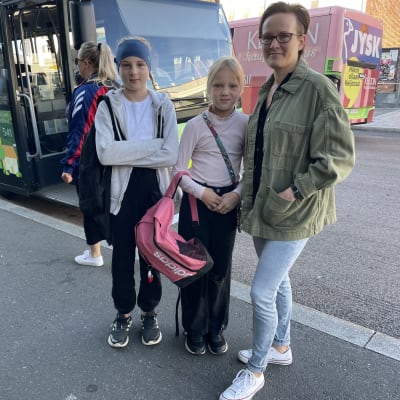 Farkkuihin ja vihreään paitaan pukeutunut nainen kahden alakouluikäisen tyttärensä kanssa bussipysäkillä Jyväskylässä.