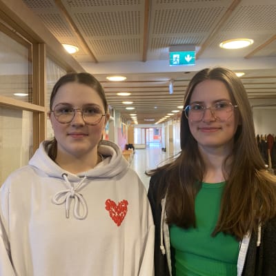 Kaksi nuorta tyttöä seisoo koulun käytävällä, katsoo kameraan ja hymyilee.