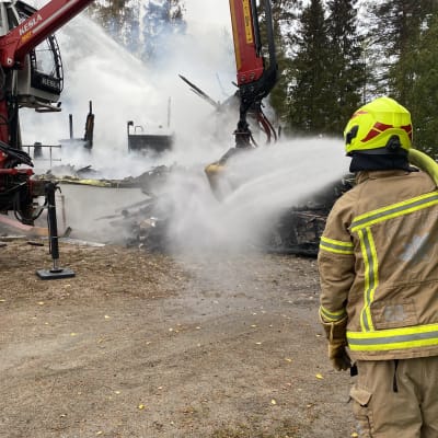 Palomies sammuttaa paloa Orimattilan Virenojalla. Taustalla maan tasalle palanut rakennus savuaa harmaana.