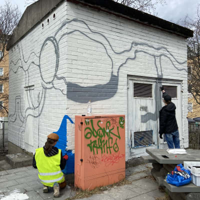 Kaksi nuorta katutaiteilijaa maalaa liikennemattoteosta Kaurialan liikennepuiston käyttämättömän rakennuksen tiiliseinään