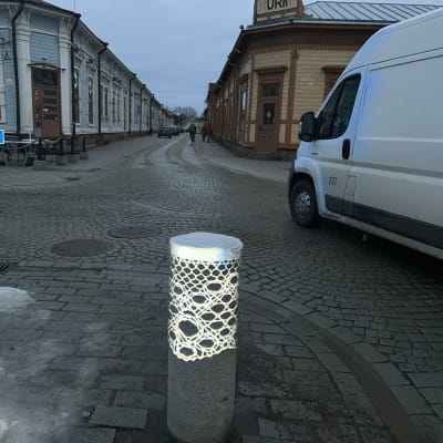 Liikenteenohjaukseen tarkoitettu tolppa, jossa on heijastinpitsi. Tolpan taustalla on harmaa auto.