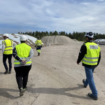 Fem personer klädda i gula västar och vita hjälmar framför stora grushögar.