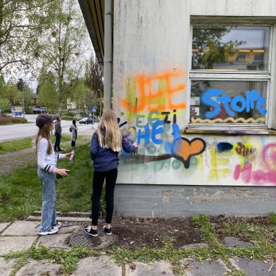 Två unga tjejer målar grafitti på skolbyggnad.