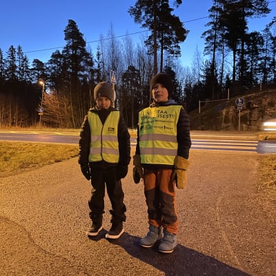 Två barn i vinterkläder och gula reflexvästar står framför ett övergångsställe över en landsväg.