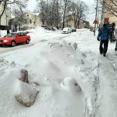 Luminen katu, punainen auto ajaa tiellä ja lumikasan vieressä kävelee talvivaatteisiin pukeutunut ihminen.