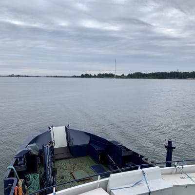 Fotot är taget på ett fartyg, på väg mot Mjölö. En del av den blåa fören på båten syns, och i bakgrunden ser man klippor som tillhör Mjölö. Det är annars mulet och grått.
