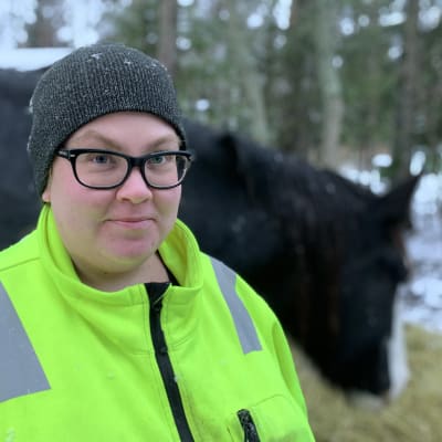 Kvinna i gul reflexjacka och mössa står utomhus i nysnö, ser in i kameran. I bakgrunden står en häst och äter hö.