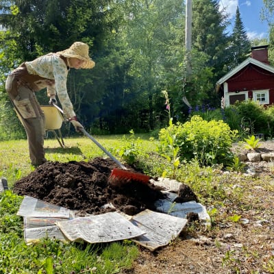 Hellehattupäinen, haalariin pukeutunut nainen (Outi Hurskainen) levittää multaa sanomalehtien päälle nurmikolla. 