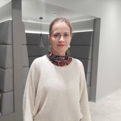 En kvinna med uppsatt ljust hår och örhängen. Står framför ett bås med soffar där man kan sitta.