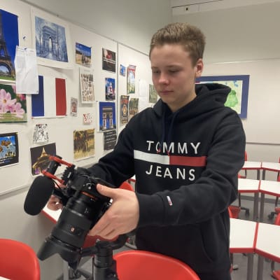 Tummansiniseen huppariin pukeutunut nuori mies seisoo koululuokassa kädessään järjestelmäkamera