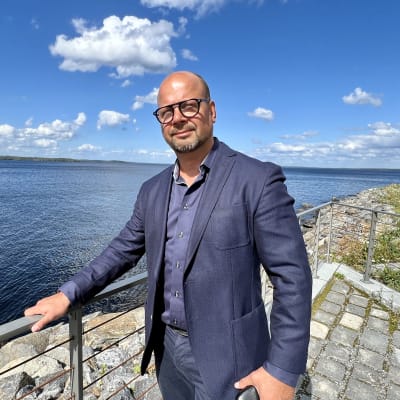 Siniseen pukuun pukeutunut silmälasipäinen mies seisoo järven rannassa.