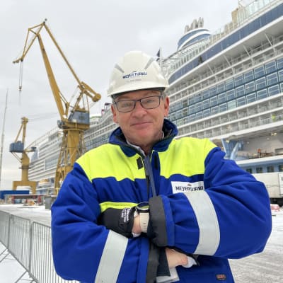 Valkokypäräinen telakkatyöntekijä seisoo ison laivan edessä ja katsoo kameraan.